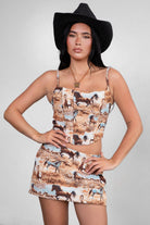 A model wearing a brown western-themed horse print crop top with adjustable shoulder straps and a square neckline. The top is paired with a matching skirt and accessorized with a wide-brimmed hat and a necklace.