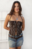 Woman wearing TIC TOC TA30581 RAELYNN embroidered western corset top with halter neckline, flame embroidery, gold studs, and lace-up front paired with a denim mini skirt