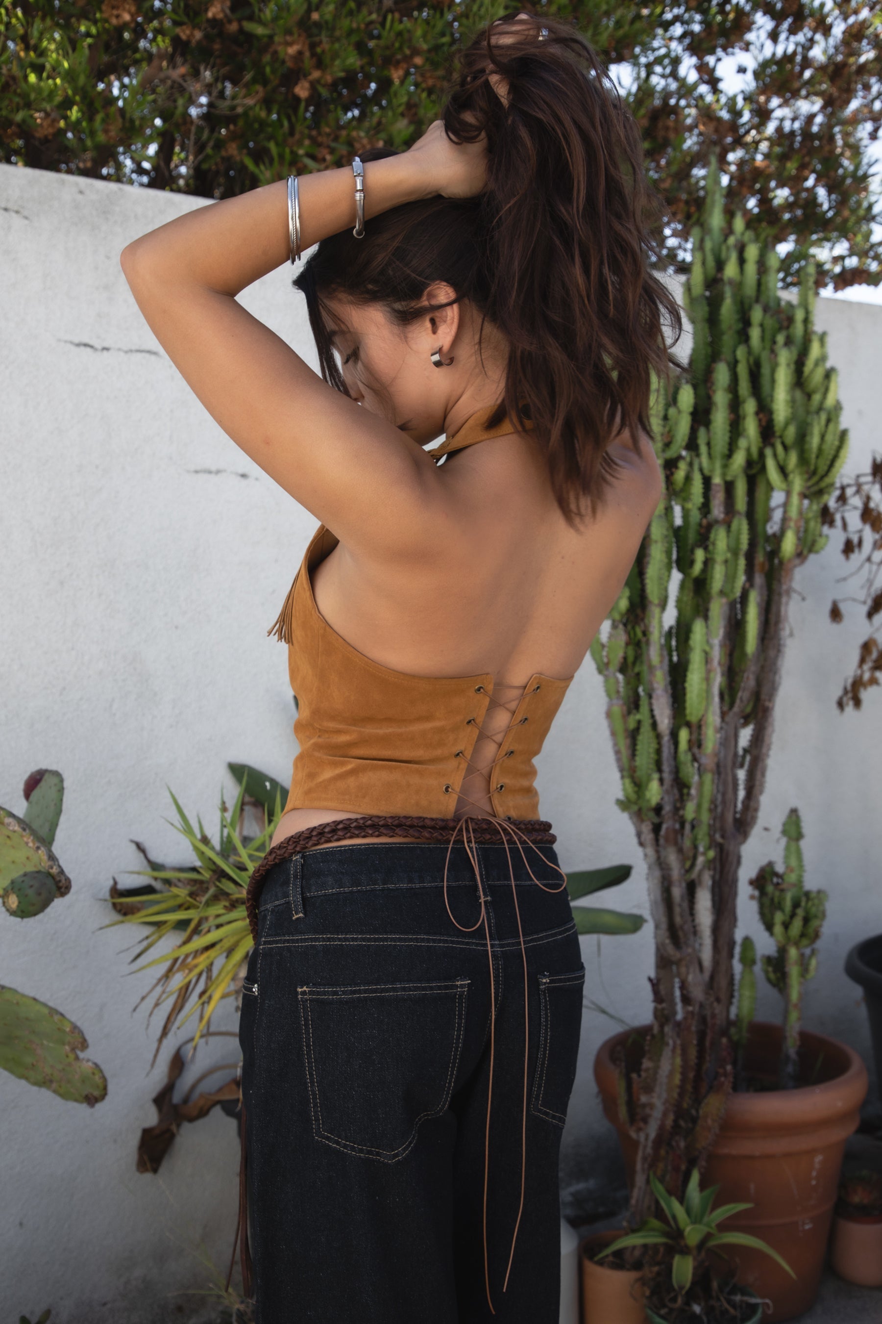 Woman wearing the TIC TOC TA31474 JOSIE tan faux suede fringe halter top featuring layered fringe neckline, lace trim tie detail, and open back with lace-up closure and dark jeans against a white wall and cacti in the background