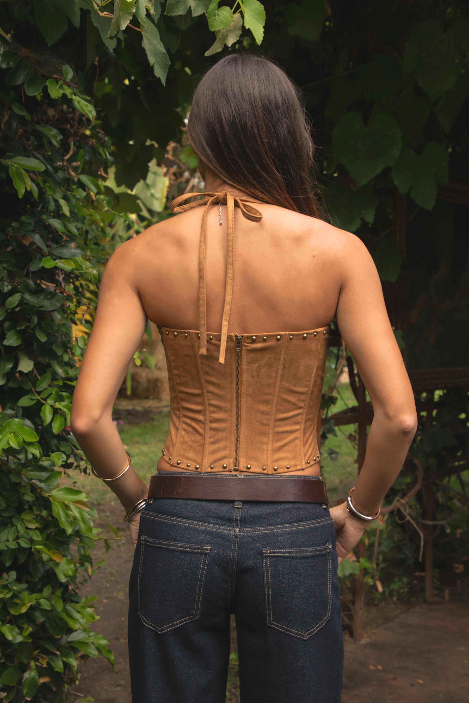 Woman wearing the TIC TOC TA31568 RIVIARA stretch suede western corset top featuring halter neckline, lace-up front, and contrast embroidery with studded trim and dark pants in an outdoor setting.