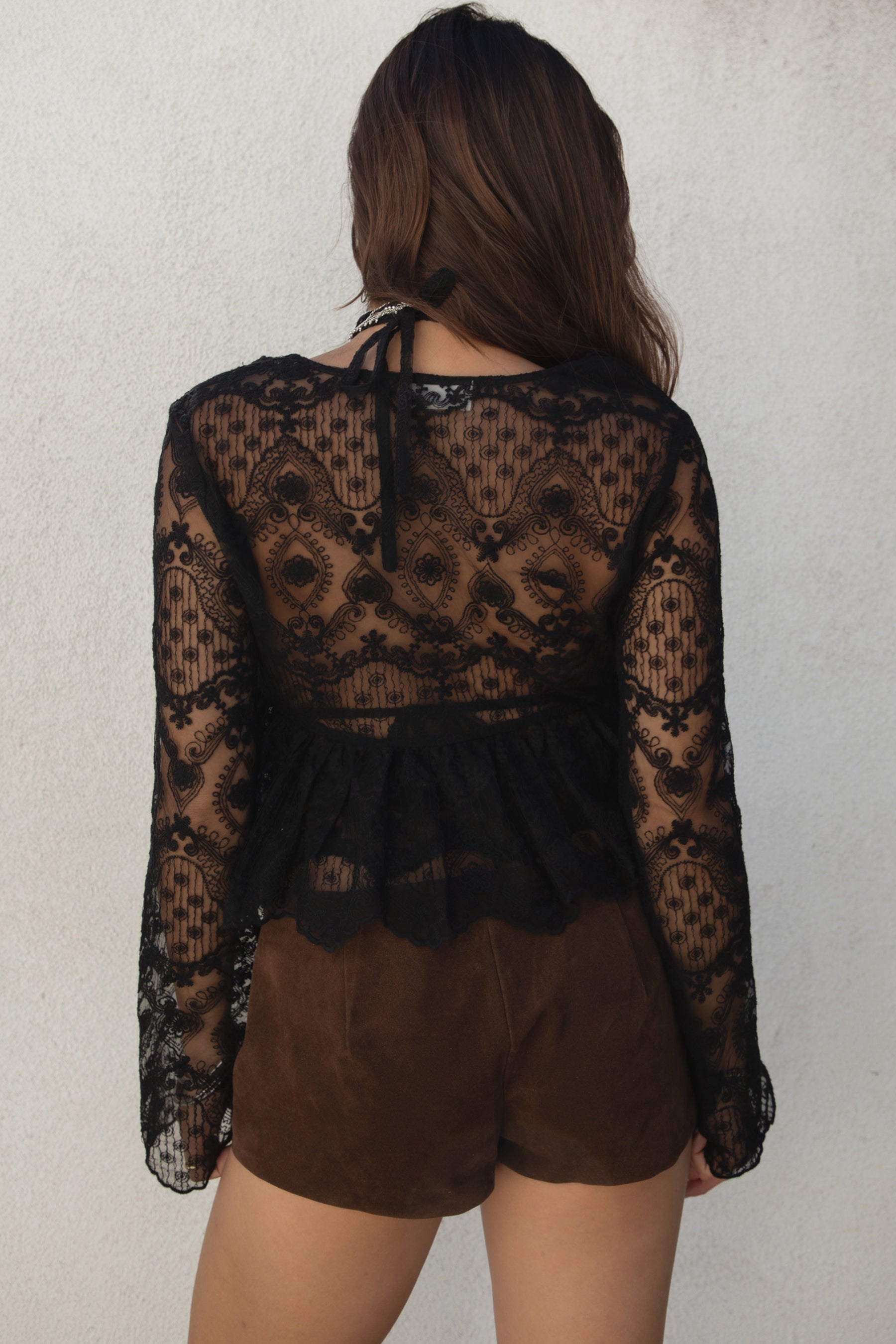 Model wearing the TIC TOC TA31608 LAREDA black crochet embroidered long sleeve top featuring a scalloped V-neckline, tie front detail, and flowy bell sleeves with suede mini shorts.