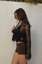 Model wearing the TIC TOC TA31608 LAREDA black crochet embroidered long sleeve top featuring a scalloped V-neckline, tie front detail, and flowy bell sleeves with suede mini shorts.