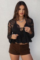 Model wearing the TIC TOC TA31608 LAREDA black crochet embroidered long sleeve top featuring a scalloped V-neckline, tie front detail, and flowy bell sleeves with suede mini shorts.