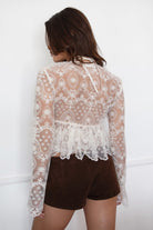 Model wearing the TIC TOC TA31608 LAREDA cream crochet embroidered long sleeve top featuring a scalloped V-neckline, tie front detail, and flowy bell sleeves with brown mini shorts. 
