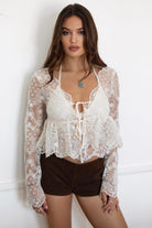 Model wearing the TIC TOC TA31608 LAREDA cream crochet embroidered long sleeve top featuring a scalloped V-neckline, tie front detail, and flowy bell sleeves with brown mini shorts. 