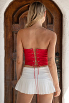 Woman wearing TIC TOC TA31611 RALLYE red stretch twill strapless top with sequin GAME DAY patch and lace-up back paired with a white skirt with brown boots in a room.