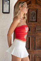 Woman wearing TIC TOC TA31611 RALLYE red stretch twill strapless top with sequin GAME DAY patch and lace-up back paired with a white skirt with brown boots in a room.