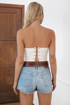 Woman wearing TIC TOC TA31611 RALLYE white stretch twill strapless top with sequin GAME DAY patch and lace-up back paired with a white skirt with brown boots in a room.