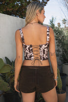 Woman wearing TIC TOC TA31648 SABLE cow print bustier corset top with adjustable straps, lace-up back, and asymmetrical hem with brown suede shorts in an outdoor setting. 