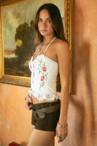 Woman wearing TIC TOC TA31649 BLYTHE white PU leather floral embroidered corset top with sweetheart neckline, lace trim, and halter tie with suede shorts in a room with a painting on the wall. 
