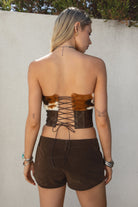 Woman wearing TIC TOC TA31671-WILDRA distressed PU leather bustier top with faux cow print fur trim and lace-up open back paired with brown suede shorts.