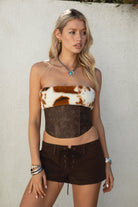 Woman wearing TIC TOC TA31671-WILDRA distressed PU leather bustier top with faux cow print fur trim and lace-up open back paired with brown suede shorts.