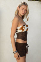 Woman wearing TIC TOC TA31671-WILDRA distressed PU leather bustier top with faux cow print fur trim and lace-up open back paired with brown suede shorts.