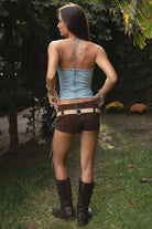 Woman wearing the TIC TOC TA31788 WRANGLER washed PU leather longline halter corset top featuring eyelet detail, lace-up cord, and back zipper in a boho-western design and brown shorts with a belt and boots, standing outdoors.
