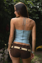 Woman wearing the TIC TOC TA31788 WRANGLER washed PU leather longline halter corset top featuring eyelet detail, lace-up cord, and back zipper in a boho-western design and brown shorts with a belt, standing outdoors.