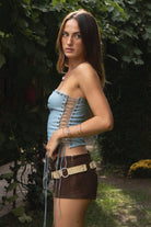 Woman wearing the TIC TOC TA31788 WRANGLER washed PU leather longline halter corset top featuring eyelet detail, lace-up cord, and back zipper in a boho-western design and brown shorts with a belt, standing outdoors.