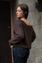 Woman wearing TIC TOC TA31803-ISARA brown oversized cable knit sweater with off-the-shoulder ribbed neckline and relaxed fit against a textured wall.