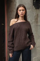 Woman wearing TIC TOC TA31803-ISARA brown oversized cable knit sweater with off-the-shoulder ribbed neckline and relaxed fit against a textured wall.