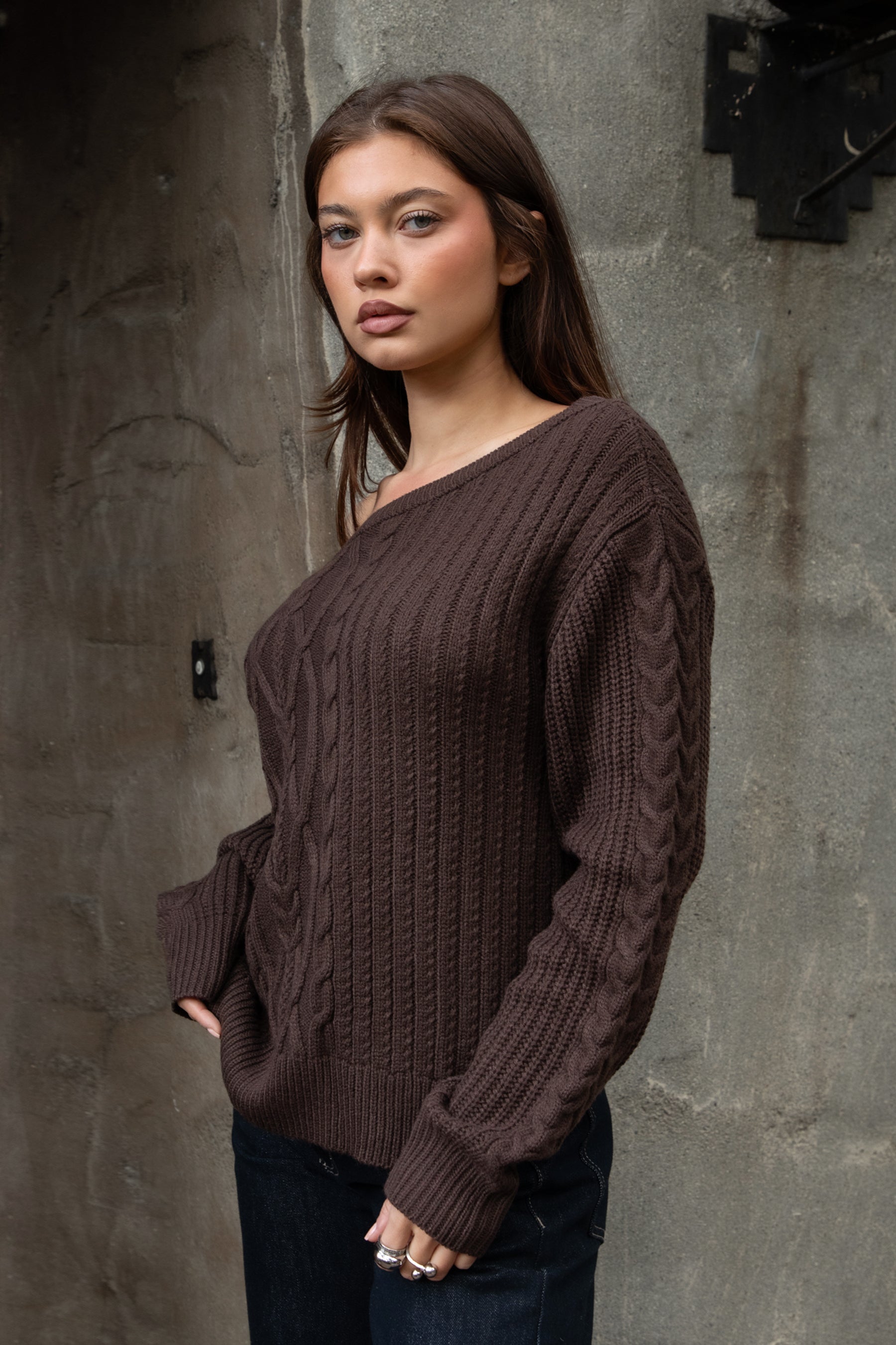 Woman wearing TIC TOC TA31803-ISARA brown oversized cable knit sweater with off-the-shoulder ribbed neckline and relaxed fit against a textured wall.