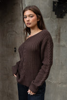 Woman wearing TIC TOC TA31803-ISARA brown oversized cable knit sweater with off-the-shoulder ribbed neckline and relaxed fit against a textured wall.