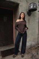 Woman wearing TIC TOC TA31803-ISARA brown oversized cable knit sweater with off-the-shoulder ribbed neckline and relaxed fit against a textured wall.