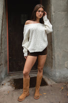 Woman wearing TIC TOC TA31803-ISARA cream oversized cable knit sweater with off-the-shoulder ribbed neckline and relaxed fit and brown shorts and tan boots against a dark background.
