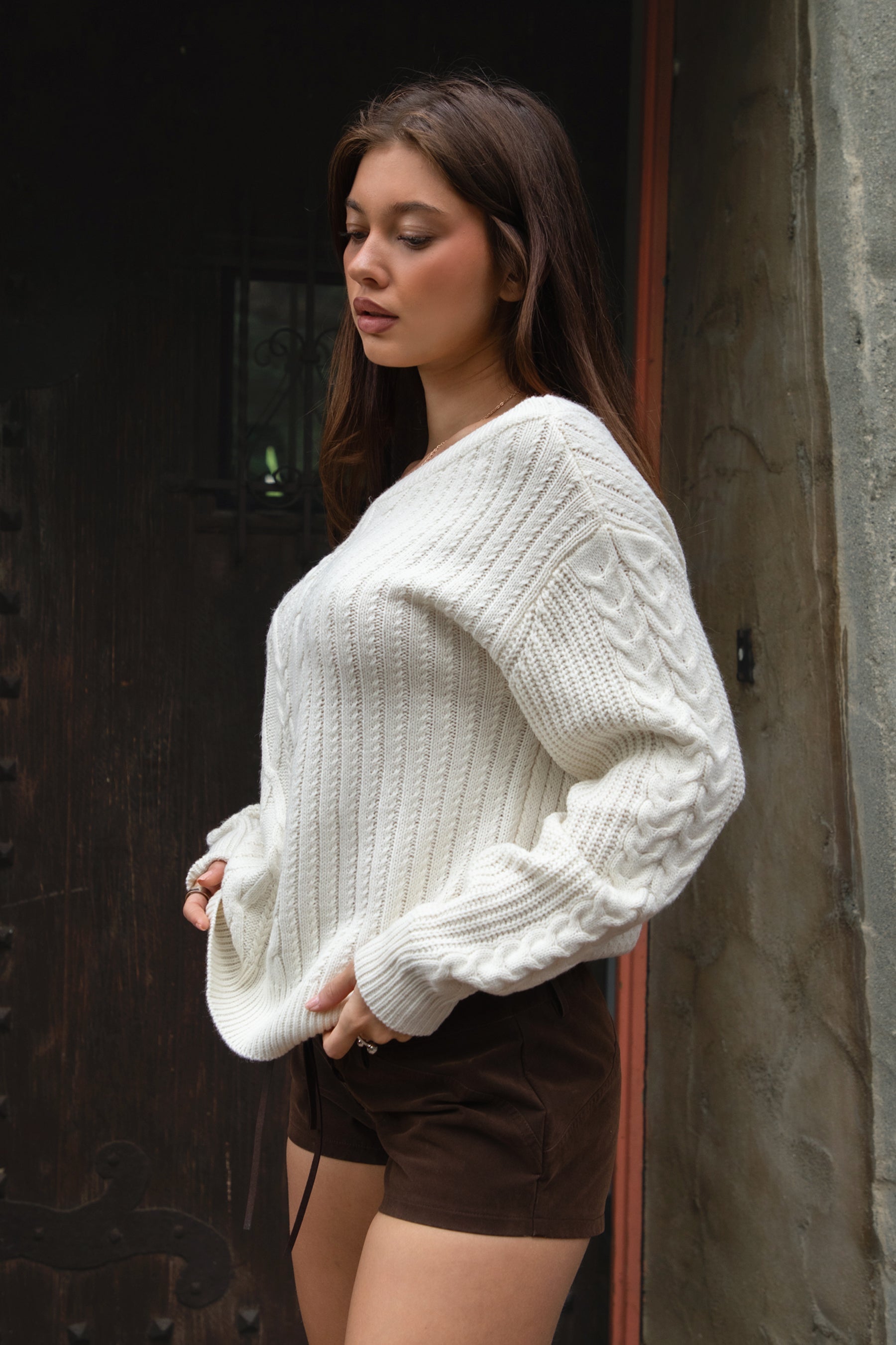 Woman wearing TIC TOC TA31803-ISARA cream oversized cable knit sweater with off-the-shoulder ribbed neckline and relaxed fit and brown shorts against a dark background.