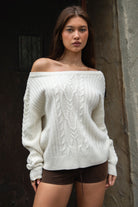 Woman wearing TIC TOC TA31803-ISARA cream oversized cable knit sweater with off-the-shoulder ribbed neckline and relaxed fit and brown shorts against a dark background.