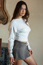 Woman wearing TIC TOC TA31843 AURELLE heather grey off the shoulder knit sweater top with dropped shoulders and relaxed long sleeves paired with a pleated brown mini skirt in an indoor setting.