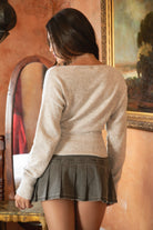 Woman wearing TIC TOC TA31843 AURELLE oatmeal off the shoulder knit sweater top with dropped shoulders and relaxed long sleeves paired with a pleated brown mini skirt in an indoor setting.
