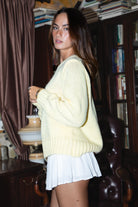 Woman wearing a light yellow sweater and white skirt in an indoor setting with books and furniture.