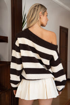 A woman wearing a TIC TOC TC30623 Maddee brown ivory rugby knit oversized sweater with an off-the-shoulder neckline and striped color block design, paired with a beige skirt, standing in a room with wooden shelves.