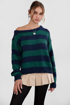 A woman wearing a TIC TOC TC30623 Maddee navy green rugby knit oversized sweater with an off-the-shoulder neckline and striped color block design, paired with a beige skirt