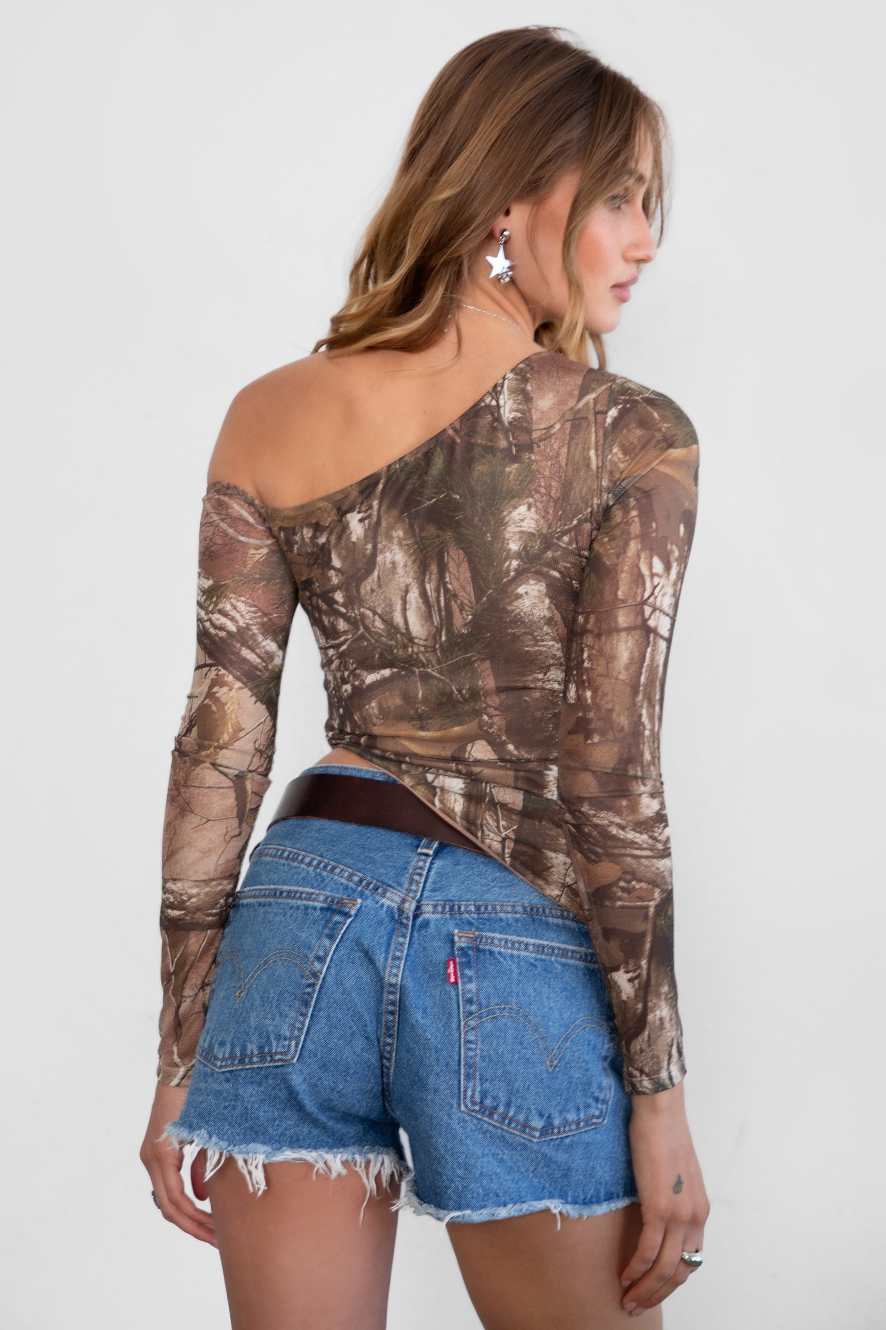 Woman wearing TIC TOC TC31252 REMIA Milk western camo print long sleeve top with asymmetrical neckline and single off-shoulder sleeve paired with blue denim shorts and western belt. 