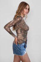 Woman wearing TIC TOC TC31252 REMIA Milk western camo print long sleeve top with asymmetrical neckline and single off-shoulder sleeve paired with blue denim shorts and western belt. 