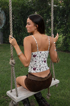 Woman wearing TIC TOC TC31307 NARIA floral mesh print cami top with plunging V neckline, satin bust detail, and adjustable straps with brown suede shorts sitting on a swing in an outdoor setting. 