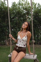 Woman wearing TIC TOC TC31307 NARIA floral mesh print cami top with plunging V neckline, satin bust detail, and adjustable straps with brown suede shorts sitting on a swing in an outdoor setting. 