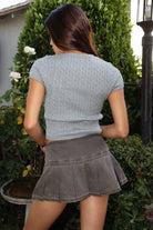 Model wearing the TIC TOC TC31437 LAURENI marled grey cable knit short sleeve top featuring a cropped silhouette, crew neckline, and textured knit design paired with a denim pleated mini skirt outdoors