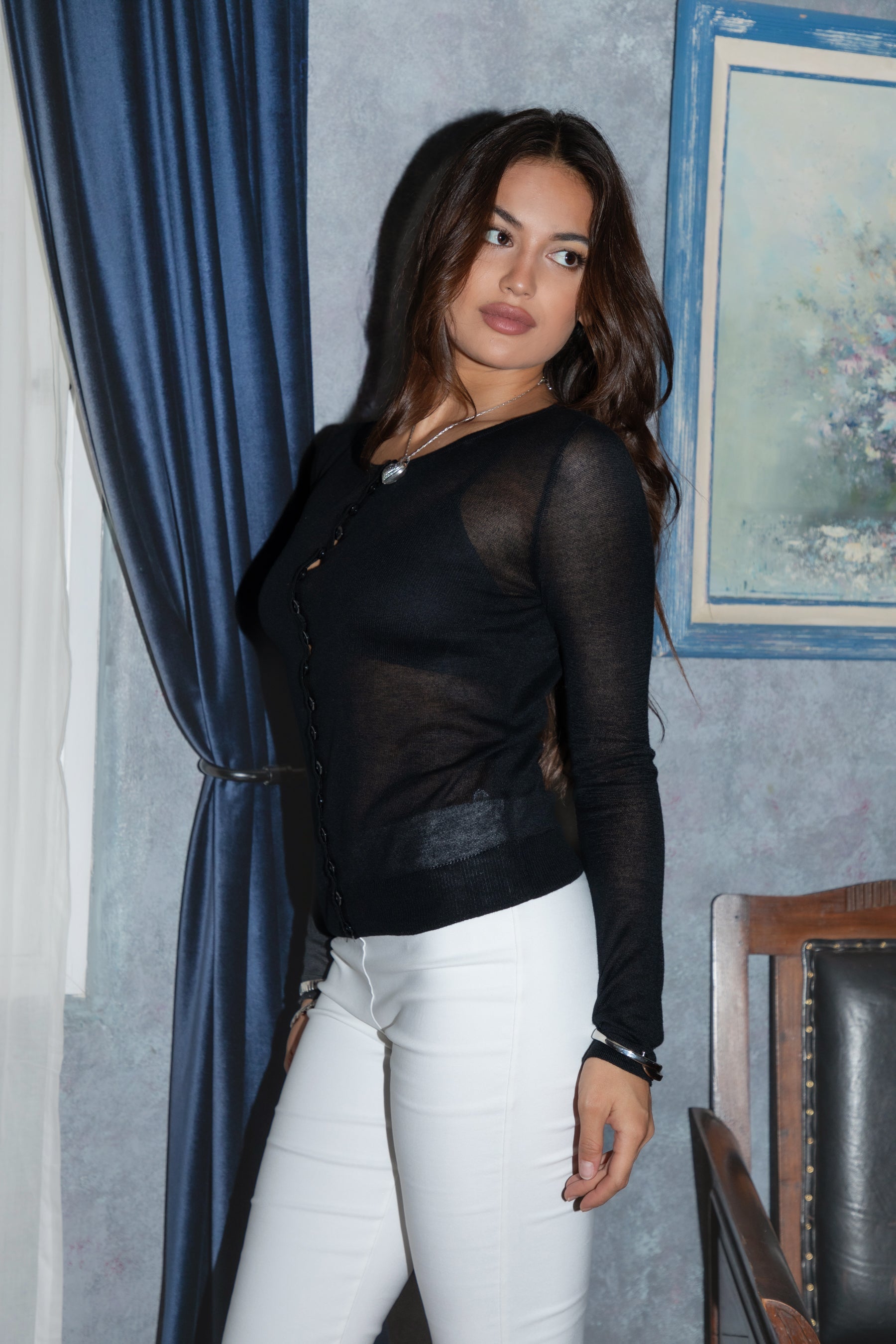 Woman wearing TIC TOC TC31469 ELISSE black  sheer knit button down long sleeve top with high scoop neckline and slim fit silhouette paired with white capri pants in an indoor setting. 