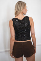 Model wearing the TIC TOC TC31578 LIIMA black sheer sequin tank top featuring a high boat neckline, sleeveless silhouette, and shimmering mesh fabric paired with brown suede shorts.
