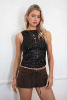 Model wearing the TIC TOC TC31578 LIIMA black sheer sequin tank top featuring a high boat neckline, sleeveless silhouette, and shimmering mesh fabric paired with brown suede shorts.