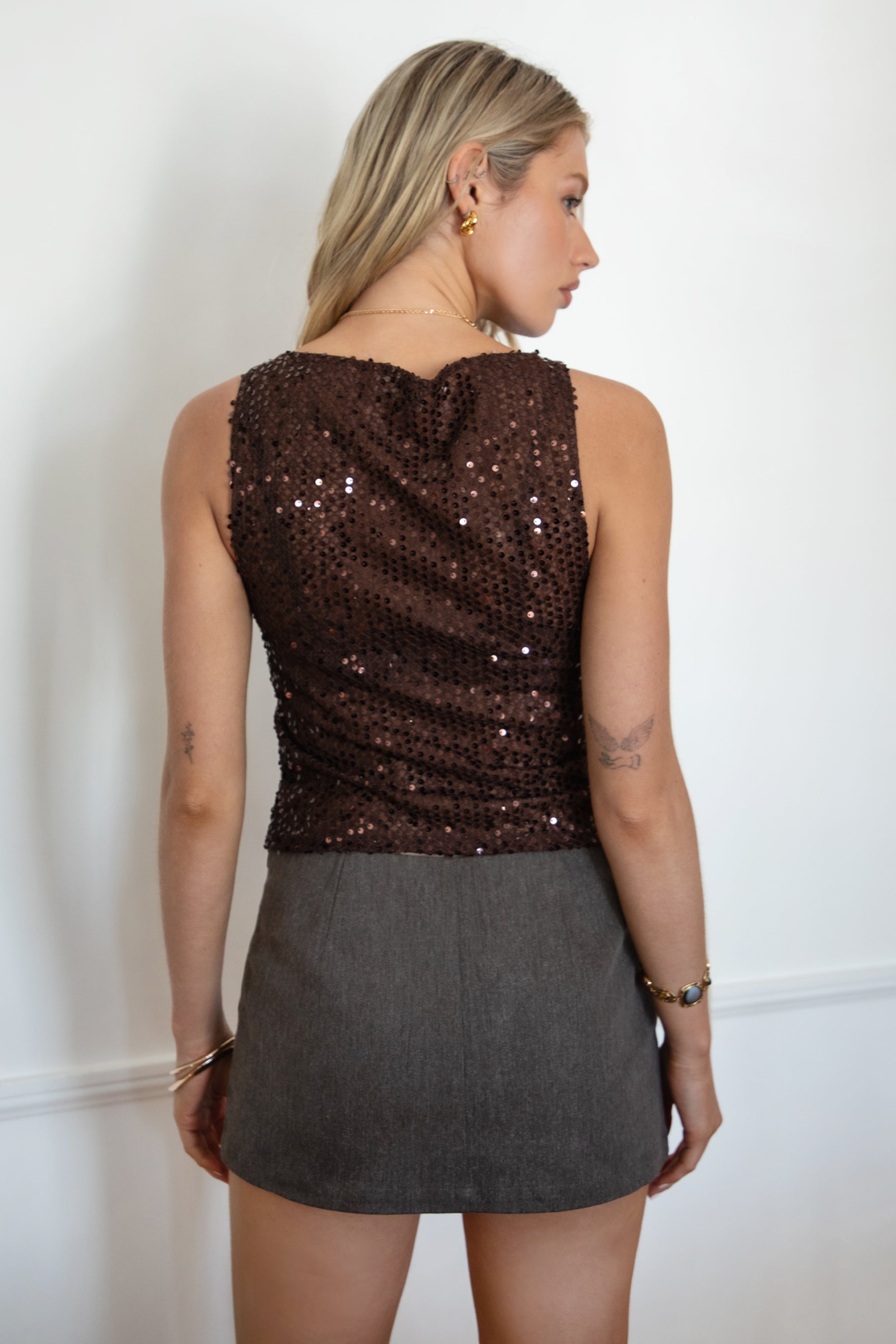 Model wearing the TIC TOC TC31578 LIIMA brown sheer sequin tank top featuring a high boat neckline, sleeveless silhouette, and shimmering mesh fabric paired with a brown denim mini skirt.