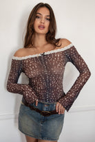 Woman wearing the TIC TOC TC31579 BAMBELLE off-the-shoulder top featuring a deer-inspired mesh print, lace trim neckline, and long bell sleeves paired with a distressed denim mini skirt