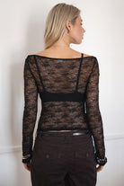 Woman wearing a TIC TOC TC31606 MIRELLE black stretch sheer lace long sleeve top featuring a wide boat neckline, fitted sleeves, and see-through lace fabric, and brown cargo pants against a white background.