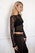 Woman wearing a TIC TOC TC31606 MIRELLE black stretch sheer lace long sleeve top featuring a wide boat neckline, fitted sleeves, and see-through lace fabric, and brown cargo pants against a white background.