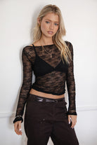 Woman wearing a TIC TOC TC31606 MIRELLE black stretch sheer lace long sleeve top featuring a wide boat neckline, fitted sleeves, and see-through lace fabric, and brown cargo pants against a white background.