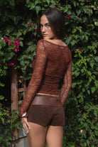 Woman wearing TIC TOC TC31606 MIRELLE brown stretch sheer lace long sleeve top featuring a wide boat neckline, fitted sleeves, and see-through lace fabric and shorts with a belt, standing against a green leafy background.