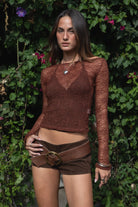 Woman wearing TIC TOC TC31606 MIRELLE brown stretch sheer lace long sleeve top featuring a wide boat neckline, fitted sleeves, and see-through lace fabric and shorts with a belt, standing against a green leafy background.