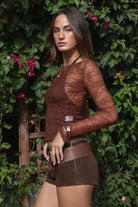 Woman wearing TIC TOC TC31606 MIRELLE brown stretch sheer lace long sleeve top featuring a wide boat neckline, fitted sleeves, and see-through lace fabric and shorts with a belt, standing against a green leafy background.