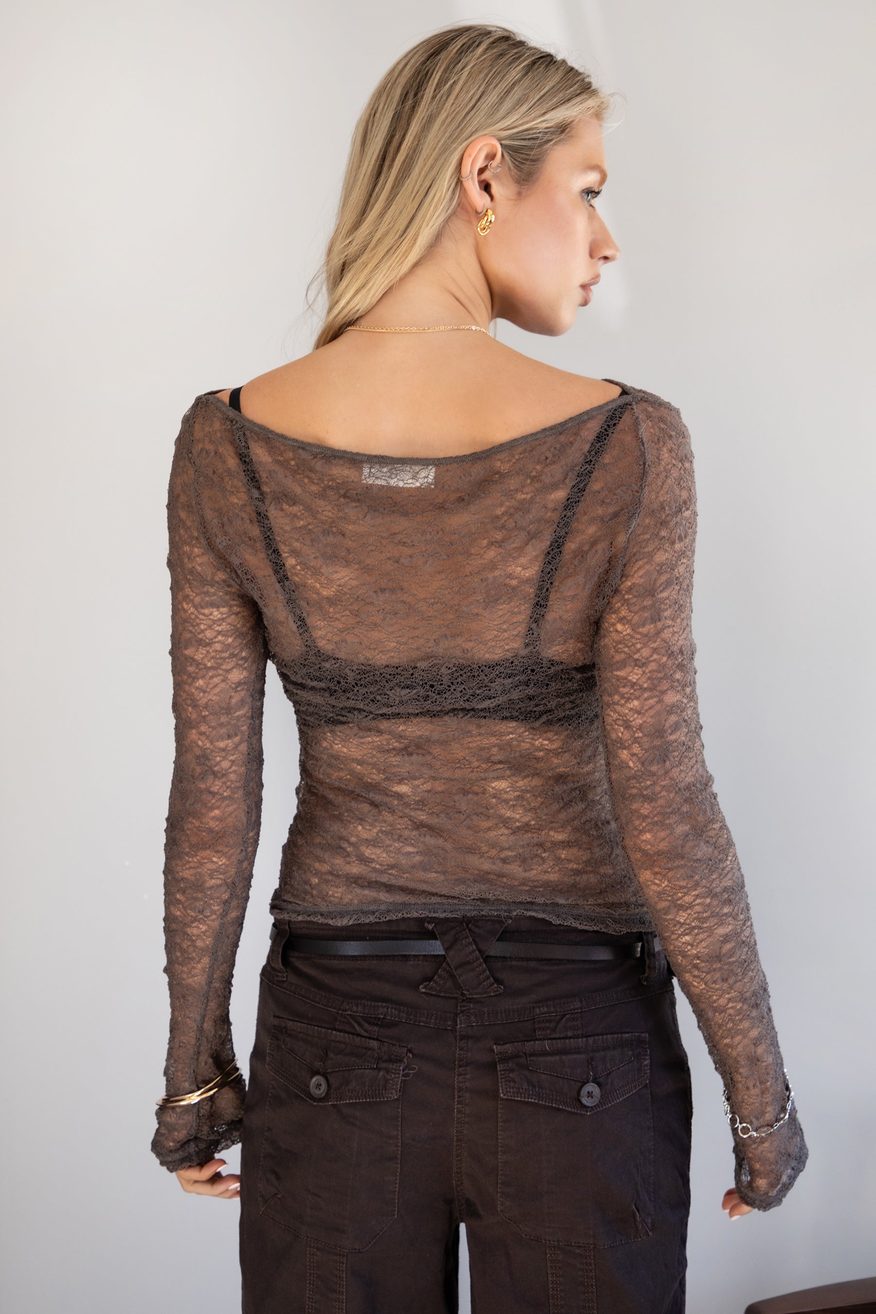 Woman wearing a TIC TOC TC31606 MIRELLE grey stretch sheer lace long sleeve top featuring a wide boat neckline, fitted sleeves, and see-through lace fabric, and brown cargo pants against a white background.